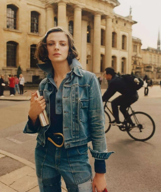 Woman in denim jacket poses in front of building, holding book, while a cyclist passes by on the street during daytime.