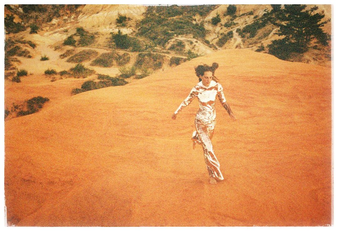 Woman in shiny metallic jumpsuit running across an orange sandy desert landscape captured on grainy film.