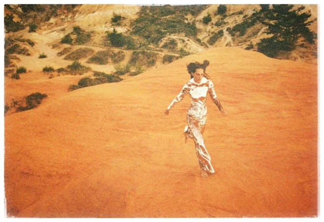 Woman in shiny metallic jumpsuit running across an orange sandy desert landscape captured on grainy film.