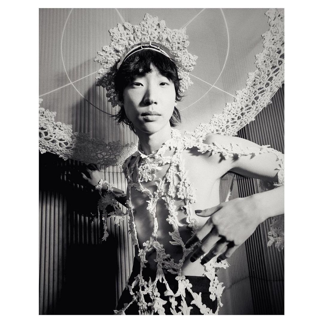 Striking black and white portrait of a person with lace headdress and shoulder covering, posing in front of a textured wall.