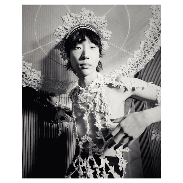 Striking black and white portrait of a person with lace headdress and shoulder covering, posing in front of a textured wall.