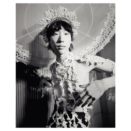 Striking black and white portrait of a person with lace headdress and shoulder covering, posing in front of a textured wall.