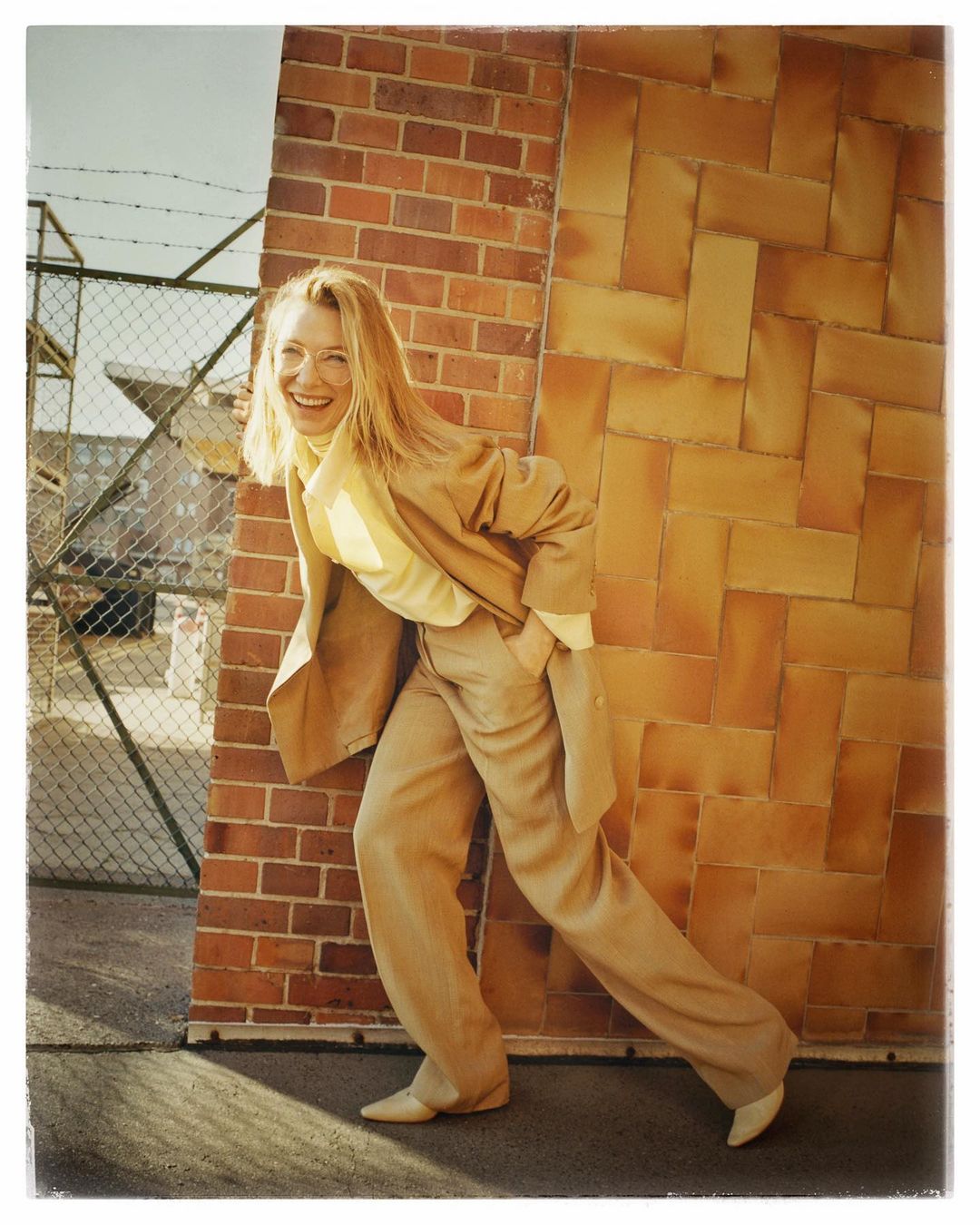 Smiling blonde woman wearing a tan trouser suit leaning against an outdoor brick wall in London