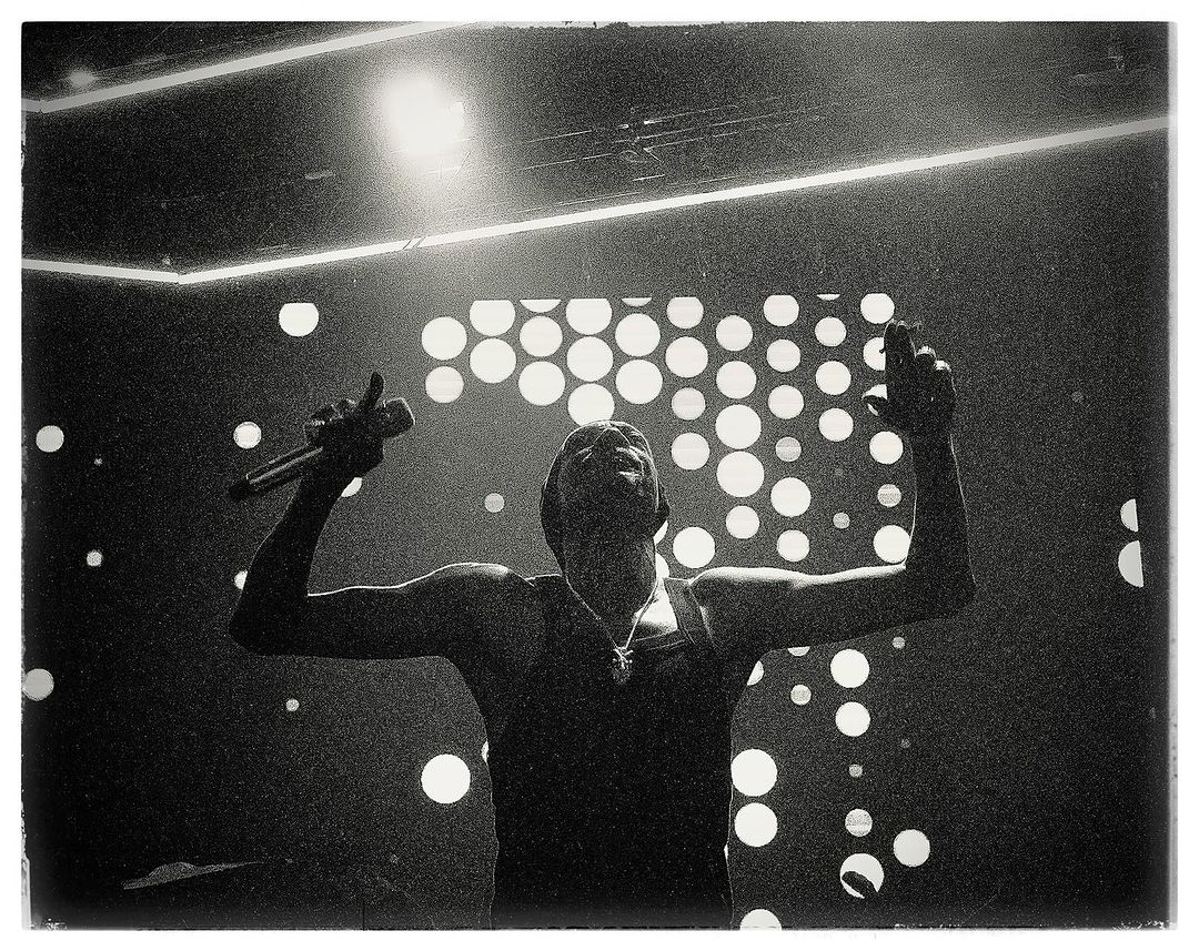 Monochrome photo of a singer performing on stage with a microphone, under bright spotlights and a backdrop of circular lights.
