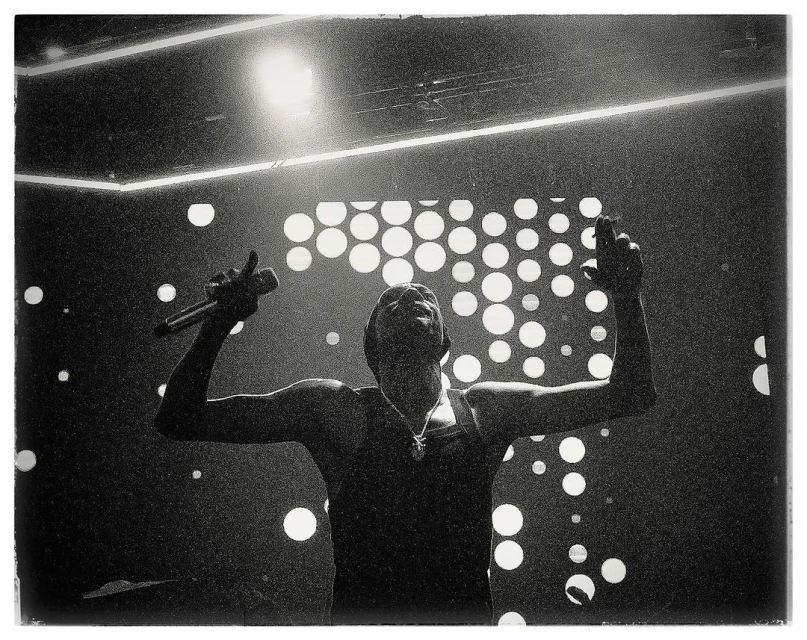 Monochrome photo of a singer performing on stage with a microphone, under bright spotlights and a backdrop of circular lights.