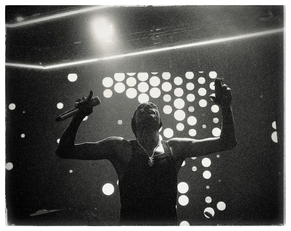 Monochrome photo of a singer performing on stage with a microphone, under bright spotlights and a backdrop of circular lights.