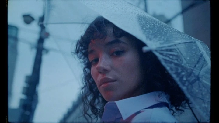 Portrait of a woman with curly hair under a clear umbrella, set against an urban background with buildings on a rainy day.