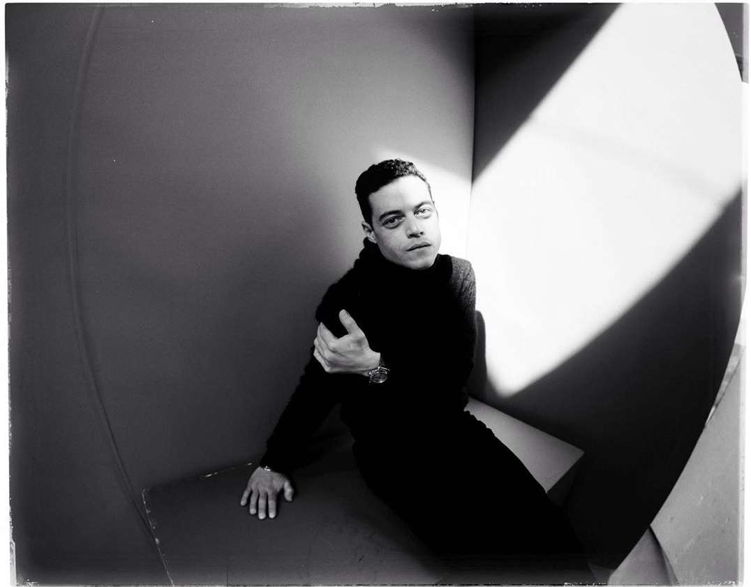 Monochrome portrait of Rami Malek indoors wearing black turtleneck, posed in a geometrically designed interior with strong light and shadow.