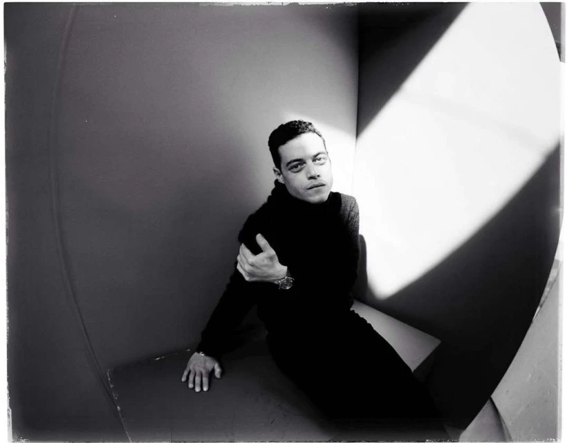 Monochrome portrait of Rami Malek indoors wearing black turtleneck, posed in a geometrically designed interior with strong light and shadow.