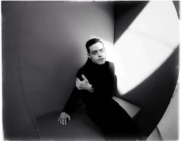 Monochrome portrait of Rami Malek indoors wearing black turtleneck, posed in a geometrically designed interior with strong light and shadow.
