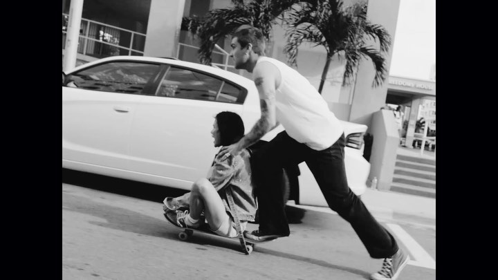 Monochrome shot of a man pushing a woman sitting on a skateboard on a city street, with a car and palm trees in the background.