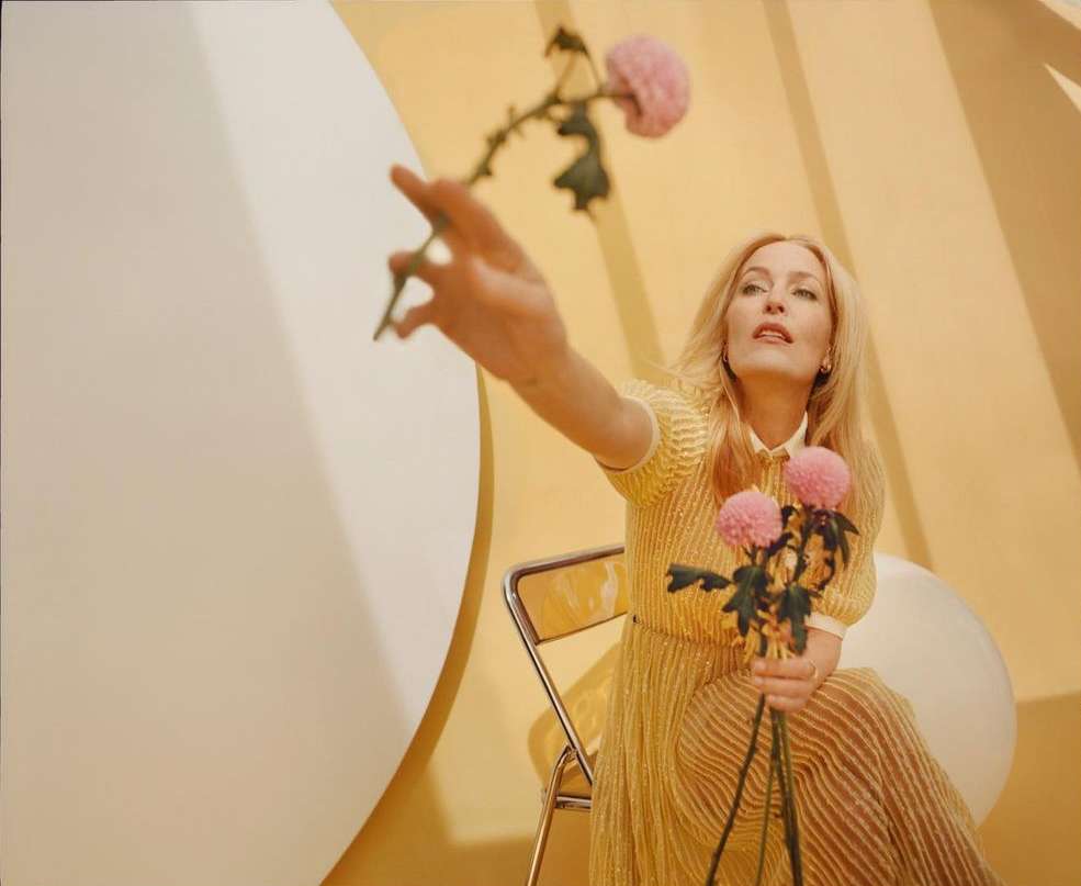 Mature blonde woman in a yellow dress holds pink flowers, reaching towards a white sphere, in a studio with a light yellow background.