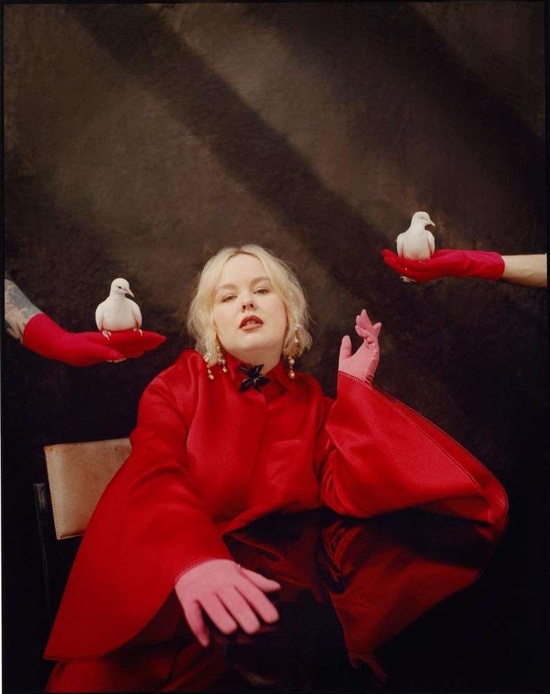 Adult woman in a red dress seated at a table with white doves on the hands of people wearing red gloves against a dark backdrop.