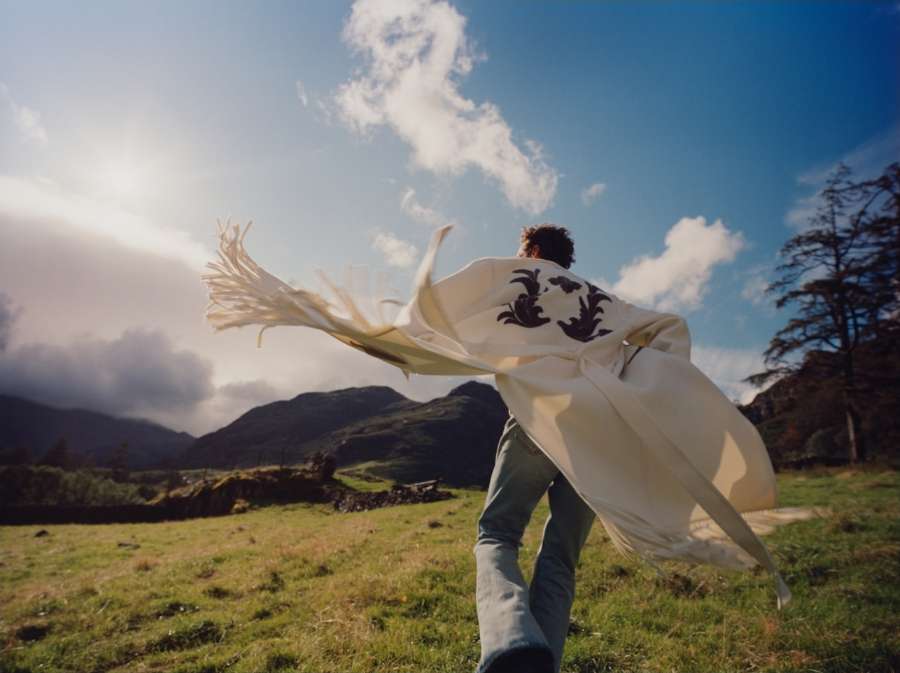 Man wearing a flowing white coat with dark embroidery running across a green meadow under a dramatic sky with mountains.