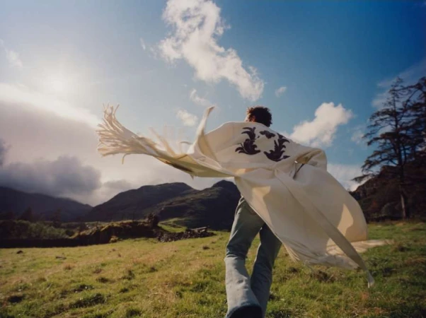 Man wearing a flowing white coat with dark embroidery running across a green meadow under a dramatic sky with mountains.
