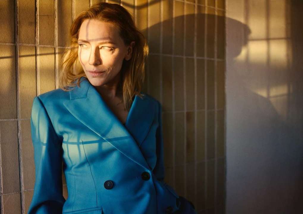 Adult female poses in a blue blazer indoors near a tiled wall with shadow patterns in a medium shot, looking to the left with sunlight.