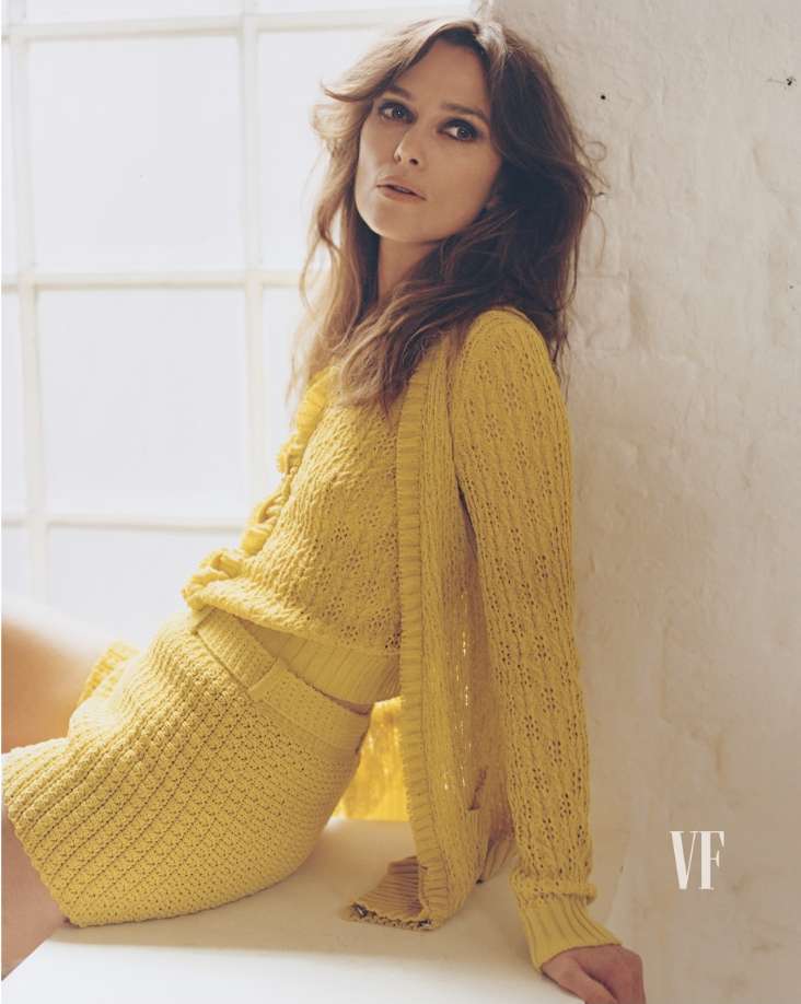 Keira Knightley poses in a yellow knit outfit with a cardigan in front of a window. She is sitting, looking slightly up at the camera.