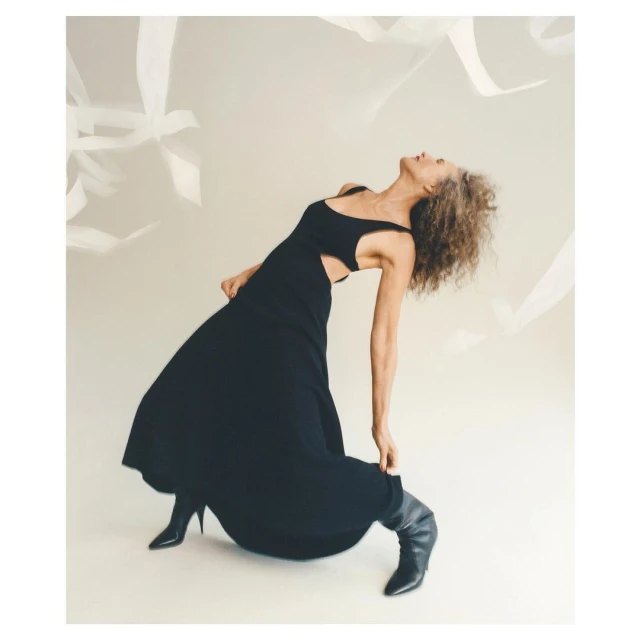 Adult woman in a black dress and black boots poses with her head tilted back in a light studio with white ribbons hanging down.