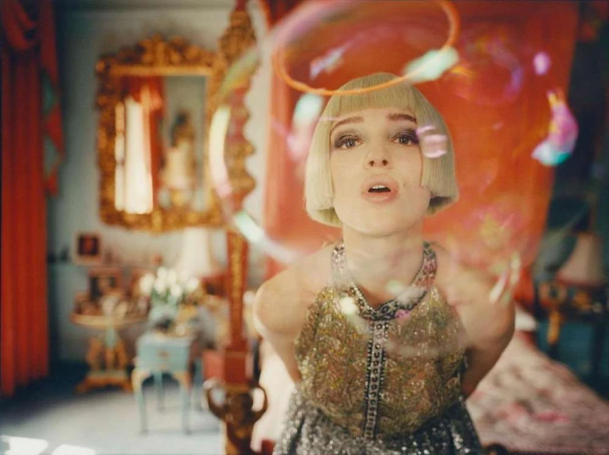 Portrait of a woman with blond bob blowing bubbles indoors. Ornate gold mirror, flowers, and red curtains in background.