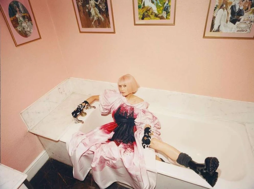 An adult woman with short pink hair and pale skin poses in a white bathtub wearing a pink dress and black boots with paintings behind her.