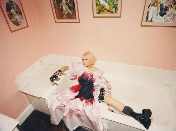 An adult woman with short pink hair and pale skin poses in a white bathtub wearing a pink dress and black boots with paintings behind her.