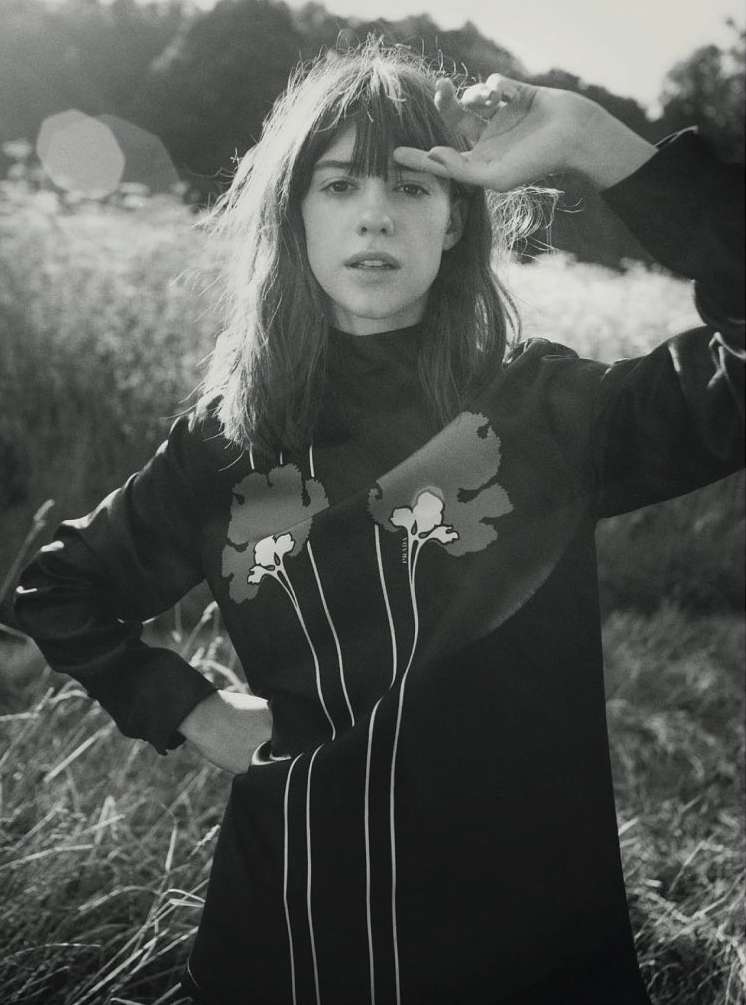 Monochrome portrait of a woman in a dress with floral designs, standing outdoors with a hand shielding her eyes from sunlight.