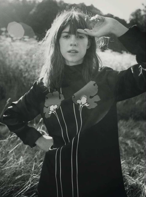 Monochrome portrait of a woman in a dress with floral designs, standing outdoors with a hand shielding her eyes from sunlight.