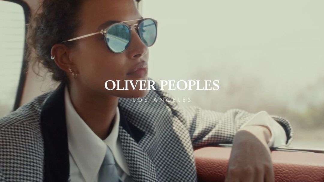 Woman in sunglasses and a patterned blazer sits inside a car with leather seats, looking off into the distance on a road trip.
