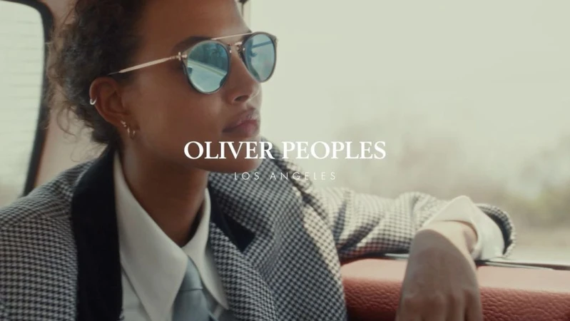 Woman in sunglasses and a patterned blazer sits inside a car with leather seats, looking off into the distance on a road trip.