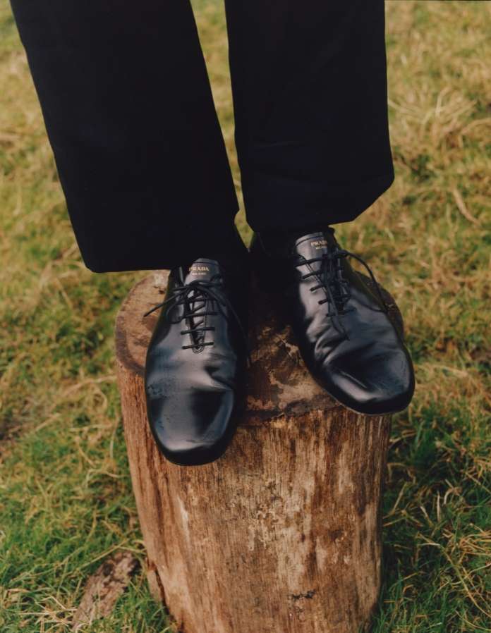 A person's black Prada shoes standing on a tree stump outdoors in a field of grass. Fashion editorial photography in natural light.