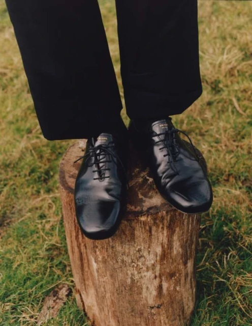 A person's black Prada shoes standing on a tree stump outdoors in a field of grass. Fashion editorial photography in natural light.