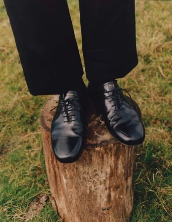 A person's black Prada shoes standing on a tree stump outdoors in a field of grass. Fashion editorial photography in natural light.