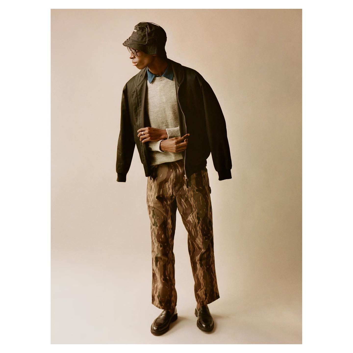 A fashionable young adult male model wears a bucket hat, jacket, sweater, and patterned pants while posing in a studio setting.