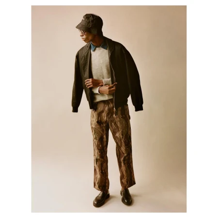 A fashionable young adult male model wears a bucket hat, jacket, sweater, and patterned pants while posing in a studio setting.