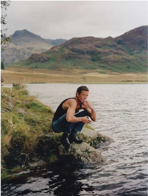 An adult male crouches on a rock next to a lake, wearing a black tank top and jeans, with mountains in the background.