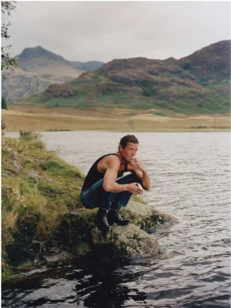 An adult male crouches on a rock next to a lake, wearing a black tank top and jeans, with mountains in the background.