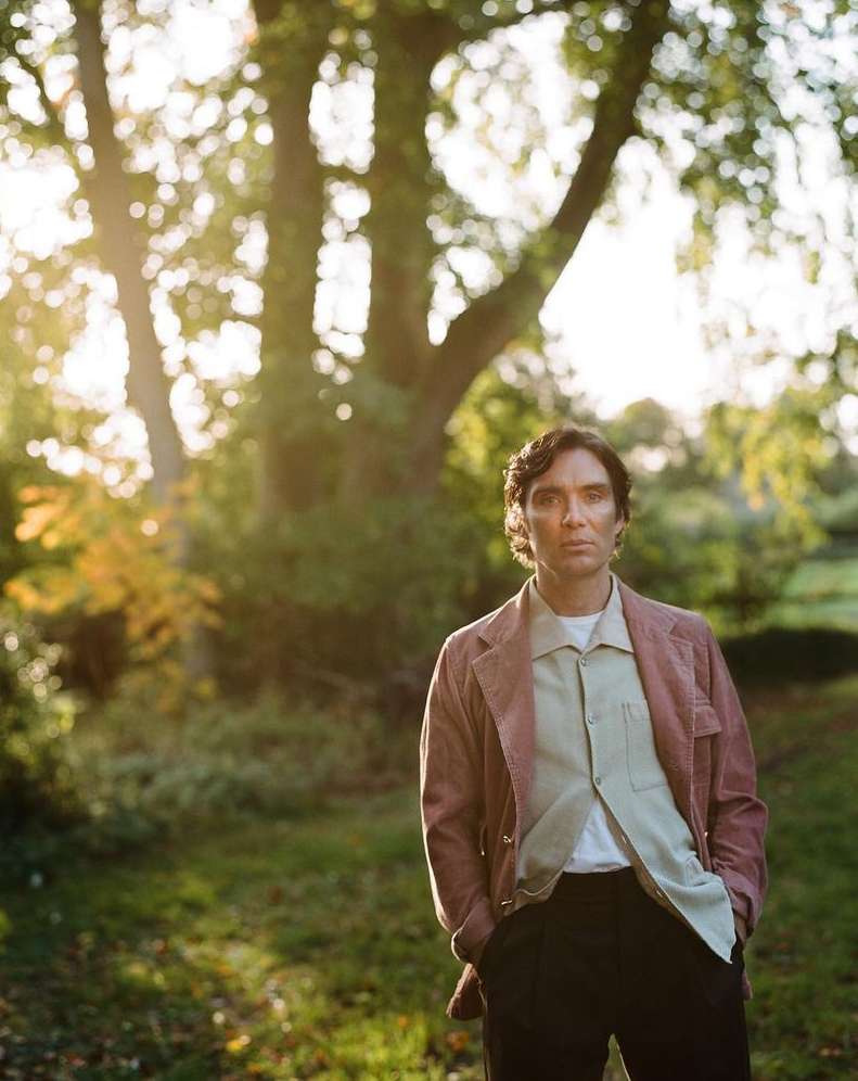 Cillian Murphy outdoors in sunlight. Adult male in a jacket and collared shirt stands in a park with large trees and green grass.