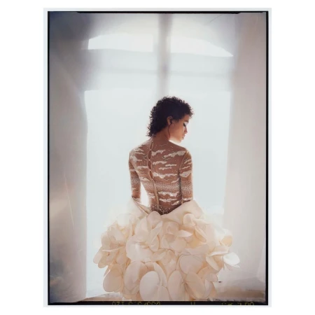 Model seen from behind wearing an ornate beaded top and voluminous petal skirt standing against a bright window.