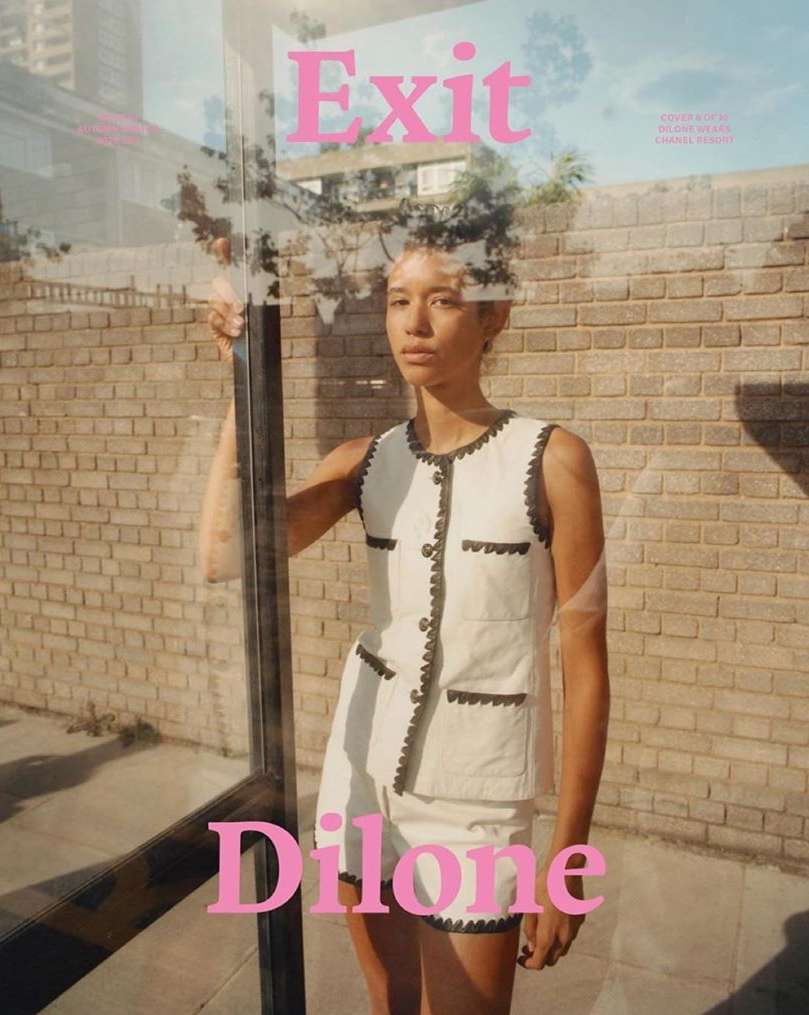 Woman in white vest and shorts posing at bus stop with transparent glass and pink text, urban fashion, outdoor shot.