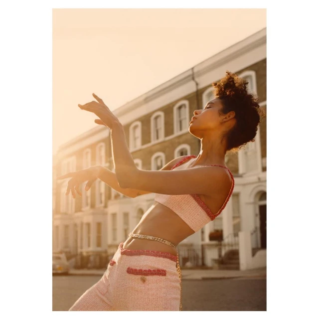 Fashion shot of woman in matching pink tweed crop top and pants, posing in front of urban street and buildings during golden hour lighting.