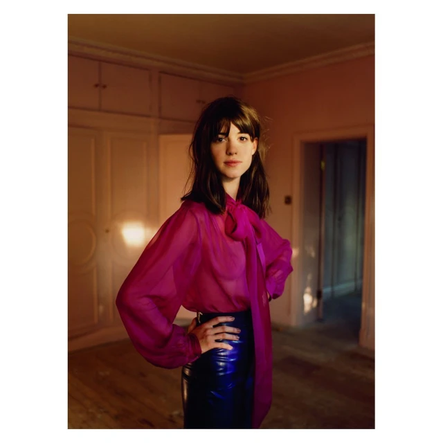 Young woman with dark hair poses in pink blouse and blue skirt in an indoor setting, looking directly at the camera with neutral expression.