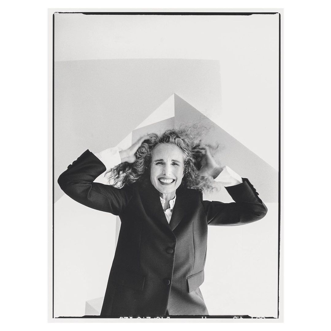 Monochrome portrait of a smiling woman with curly hair wearing a dark suit jacket against a white studio background.