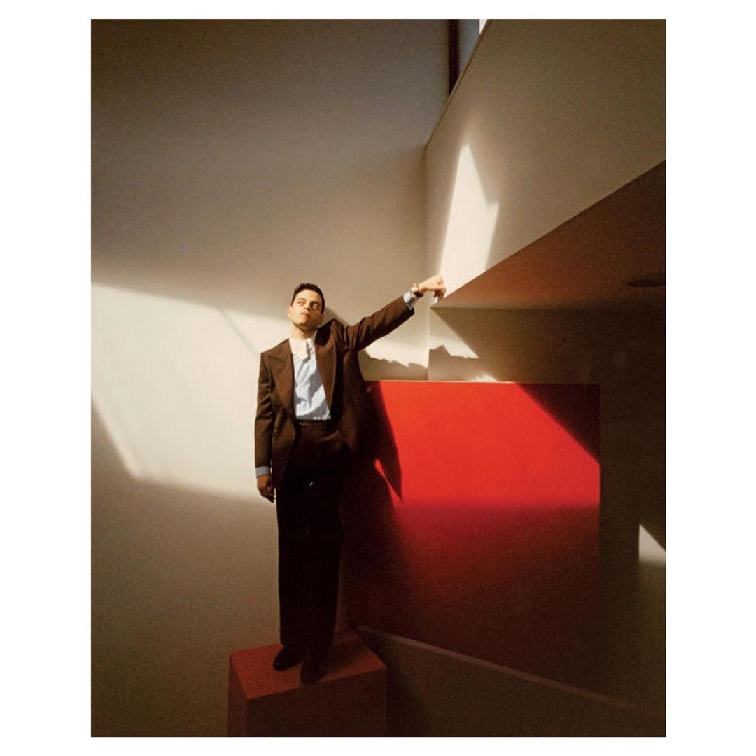 Portrait of a man in a brown suit, posing on red geometric shapes against a white wall with shadows, adding a surreal touch.