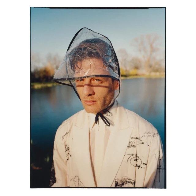 A man outdoors with a clear plastic hood, wearing a white suit with sketches; light blue sky and water in background, eye-level portrait.