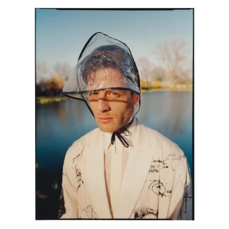 A man outdoors with a clear plastic hood, wearing a white suit with sketches; light blue sky and water in background, eye-level portrait.