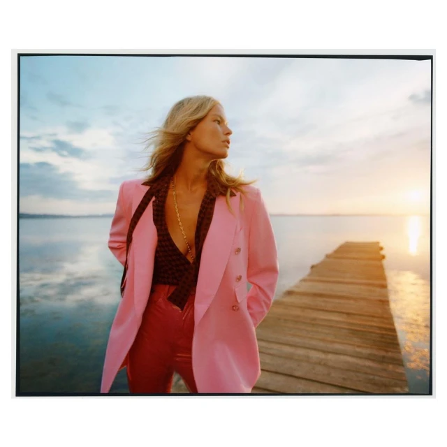 Woman in pink blazer poses on dock overlooking water at sunset. Fashion shot with a model in stylish attire enjoying a serene waterfront view.