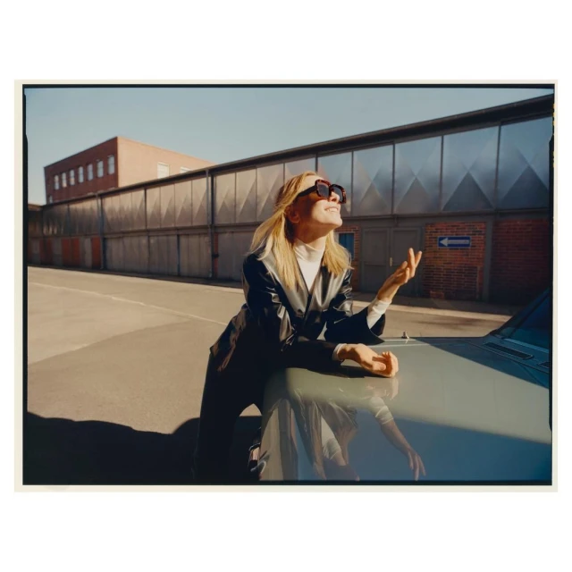 A blonde woman in a black jacket and sunglasses leans on a grey car under a clear sky, with industrial buildings in the background.