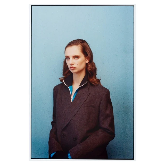 Portrait of a young woman with auburn hair wearing a gray double-breasted blazer and a turquoise top against a blue textured wall.