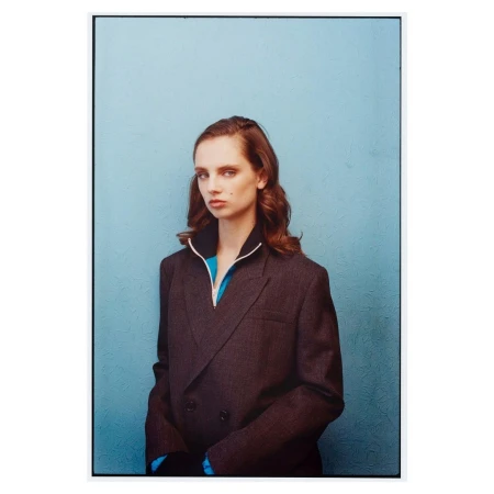 Portrait of a young woman with auburn hair wearing a gray double-breasted blazer and a turquoise top against a blue textured wall.