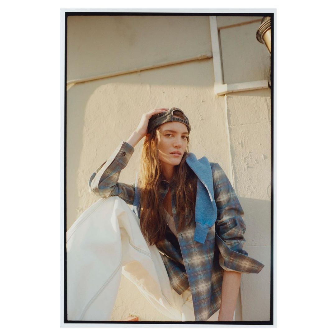 Young woman poses in plaid shirt and baseball cap against stucco wall in outdoor fashion shot, bathed in soft, diffused sunlight.